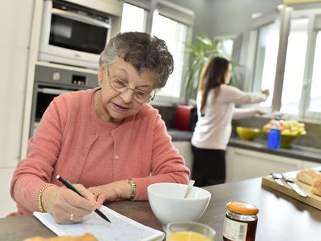 An elderly woman sitting at a desk, writing on a piece of paper with a pen.