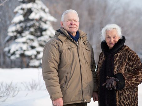 An elderly couple standing in the snow, holding hands and smiling.